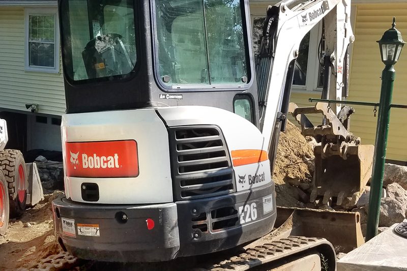 Miniescavadora compacta de rastos junto a terra escavada, ao lado de uma moradia